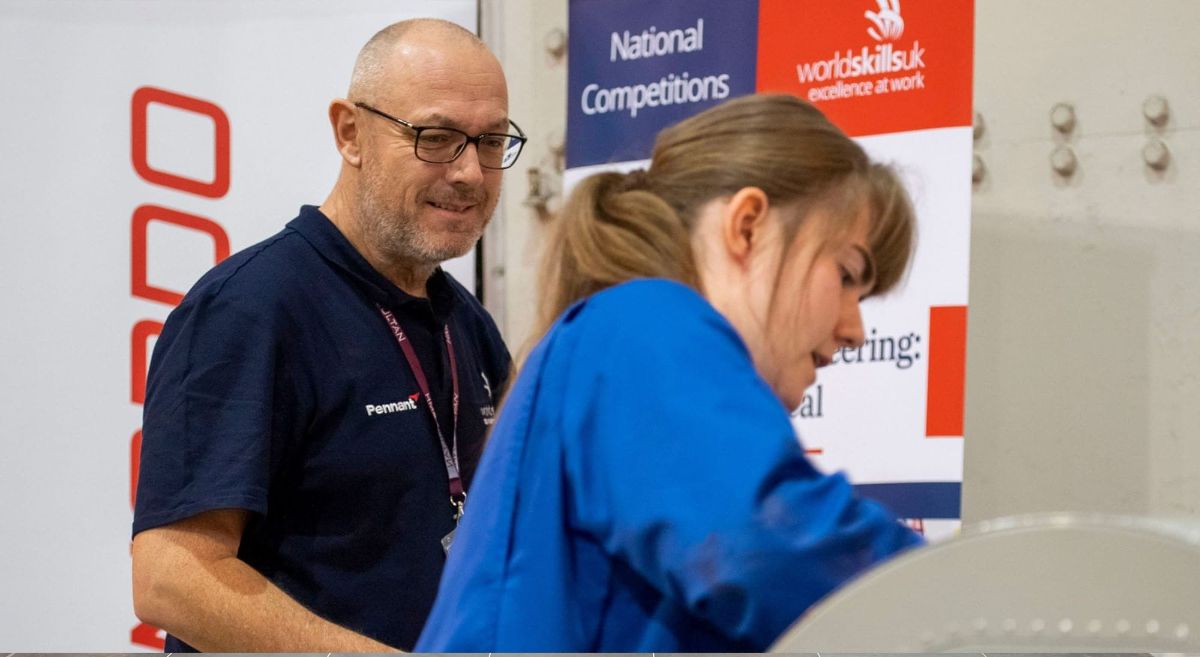 Man in WorldSkillsUK navy teeshirt looking on as female student in blue overalls works on machine.